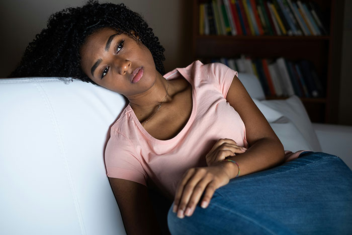 A woman experiencing grief, reclined on a white sofa, wearing a pink shirt, with bookshelves in the background.