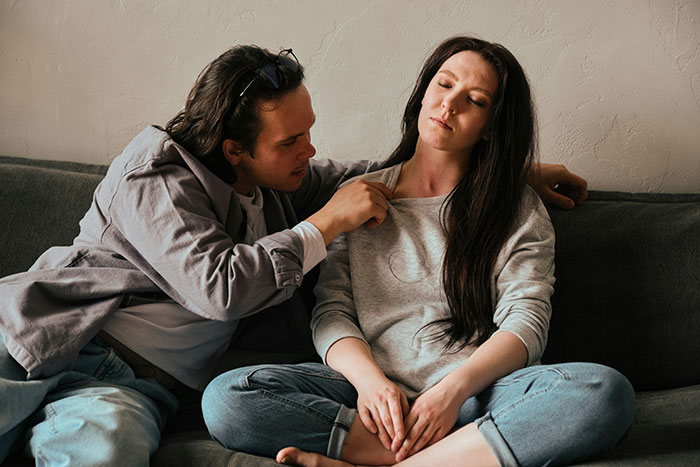 A man comforting a woman on a couch, both exhibiting emotions related to grief.