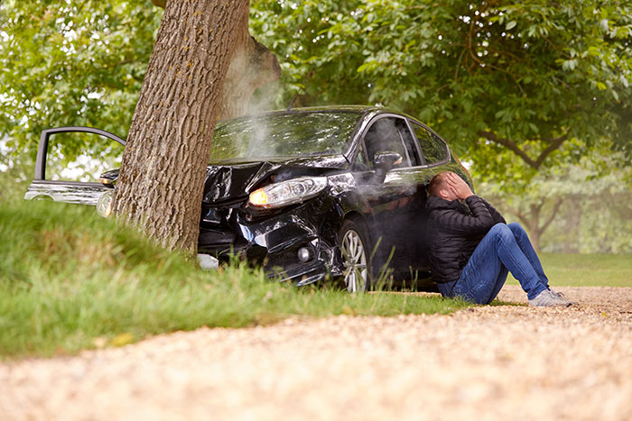 Car crashed into tree with a man sitting on the ground distressed, illustrating how to ruin your entire life.