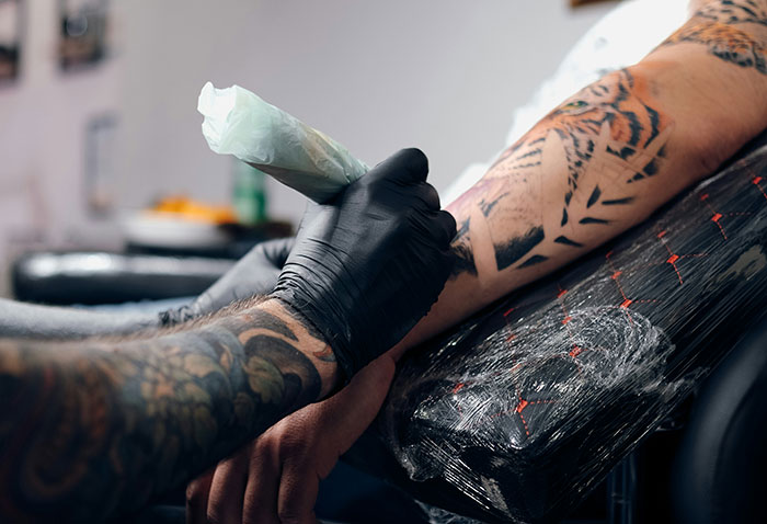 Tattoo artist wearing gloves applies ink to heavily tattooed arm, illustrating stories on how to ruin your entire life.