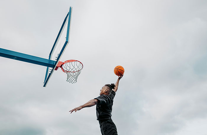 Young man jumping to slam dunk a basketball on outdoor court under cloudy sky, symbolizing how to ruin your entire life mistakes.