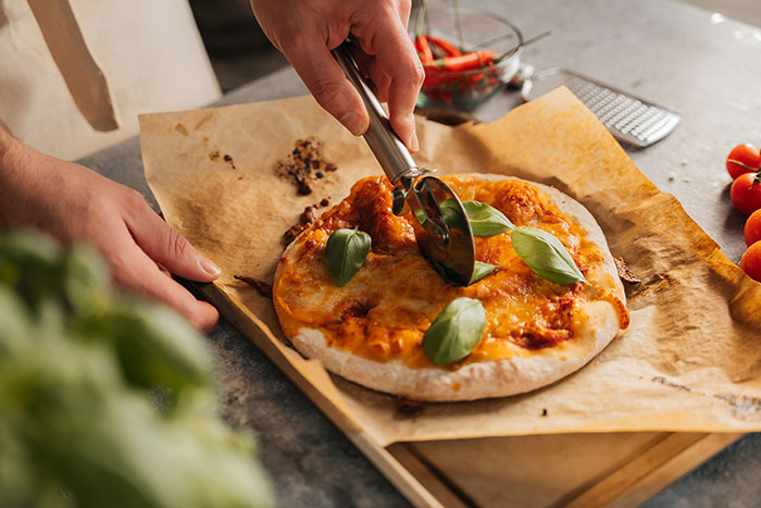 Person slicing homemade pizza on parchment, demonstrating frugality in the kitchen with fresh basil and simple ingredients.