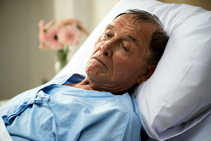 Elderly man in hospital bed, appearing thoughtful, wearing a blue gown, possibly reflecting on frugality decisions.