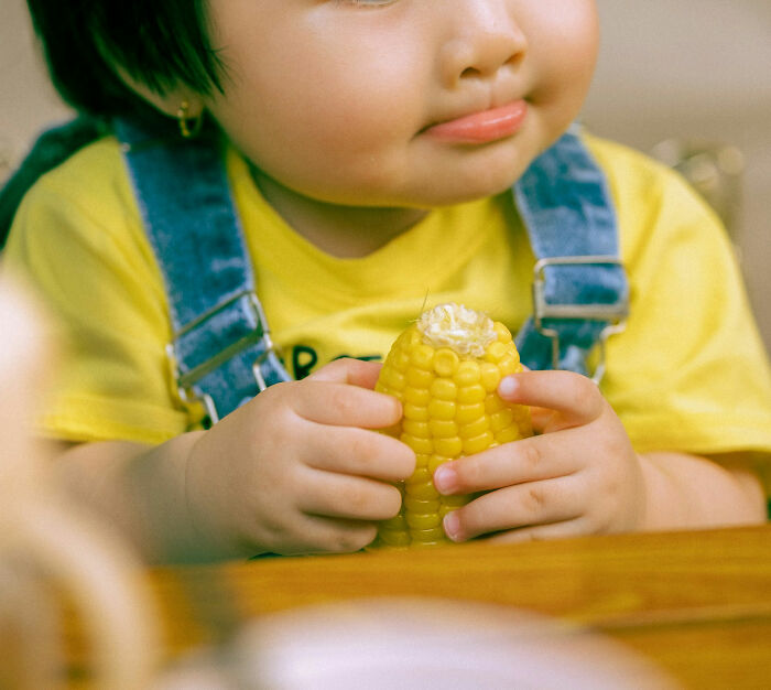 Child eating corn on the cob, wearing denim overalls and a yellow shirt, showcasing a typical family moment.