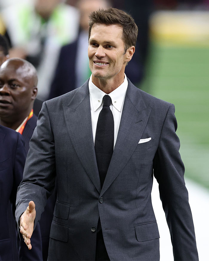 Man in a dark suit and tie at Super Bowl 2025, smiling and extending hand, embodying a head-turning look.