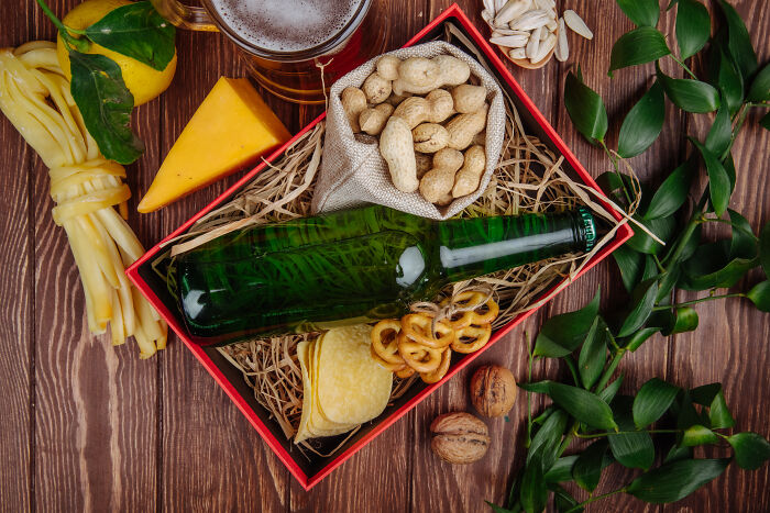 Gift ideas including a bottle, peanuts, and cheese in a decorative box on a wooden table with green leaves.