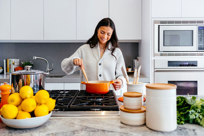 Home cook smiling while stirring food on a stove in a bright kitchen, surrounded by cooking utensils and fresh ingredients. Home cook smiling while stirring food on a stove in a bright kitchen, surrounded by cooking utensils and fresh ingredients.