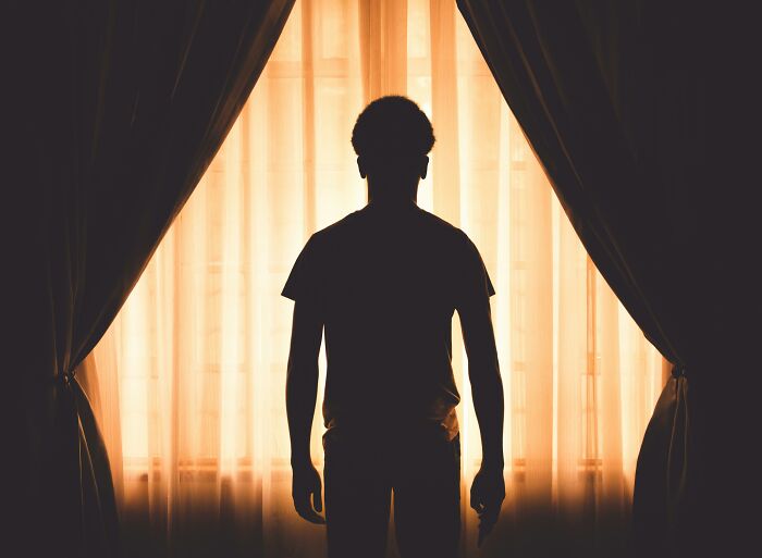 A silhouette of a person stands by a window, framed by curtains, depicting a sense of solitude and trauma.