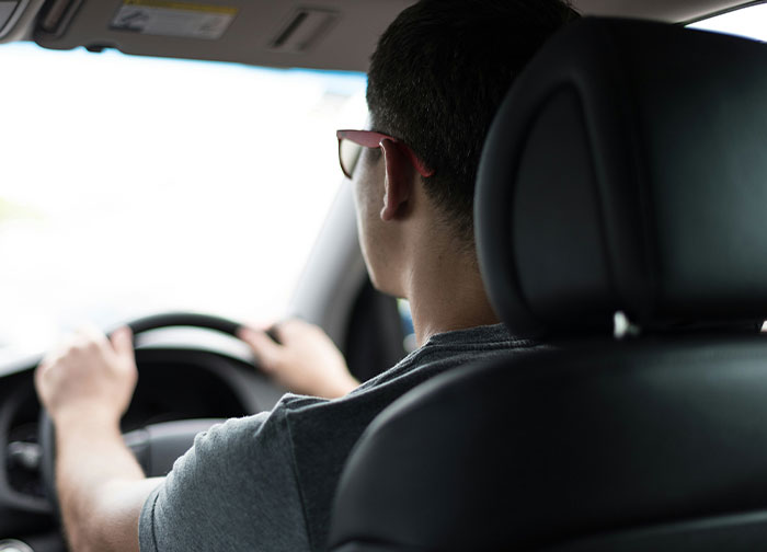 Person driving a car, wearing sunglasses, viewed from the backseat, reflecting on questions about homeland.