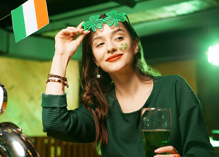 Woman in green outfit with shamrock glasses holding a beer, celebrating her homeland.