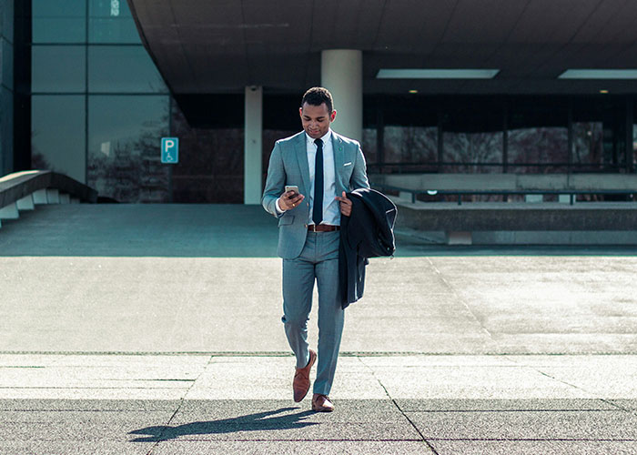A man in a suit walking outdoors, checking his phone, staying active to reach 10K steps a day.