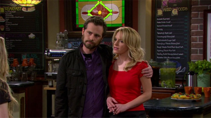 TV couple standing in a café, lacking chemistry, man in dark jacket with arm around woman in red top.