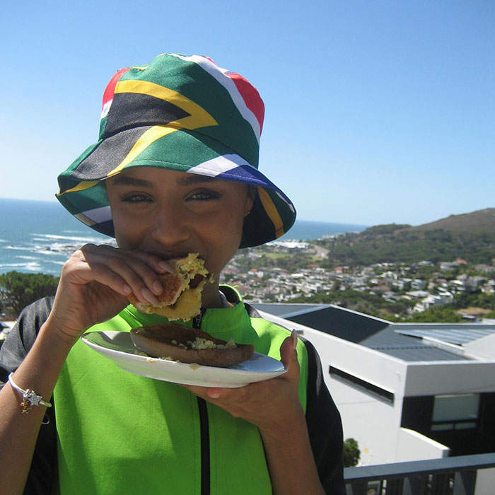 Tyla in a South African flag hat eating outside with a scenic background, reflecting Sharleen and Sherwin Seethal's prominence.