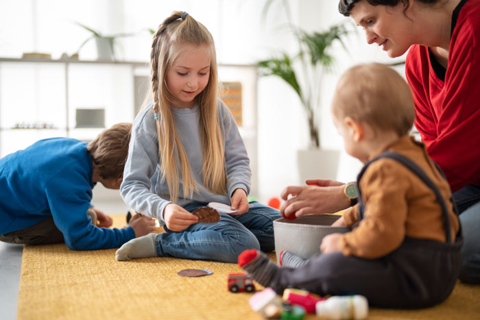 A woman interacting with young children on vacation, surrounded by toys and a play mat indoors.