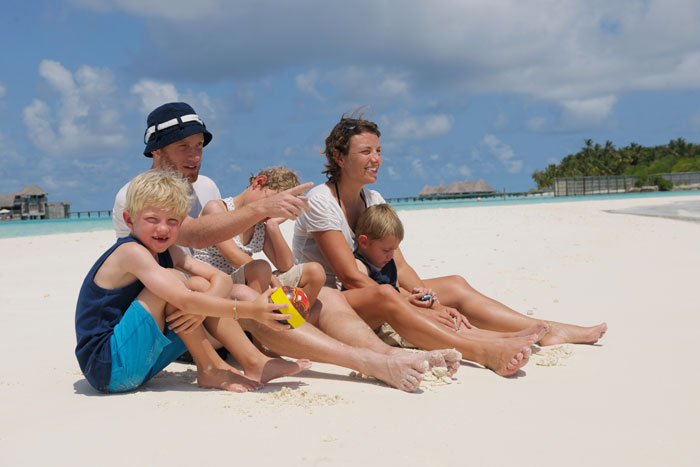 Family enjoying a beach vacation, two adults sitting with three children on the sand under a clear blue sky.