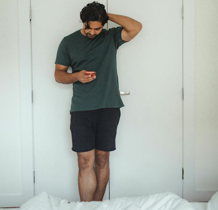 A person with prominent nose relaxedly looks at phone in casual home setting, wearing a green shirt and black shorts.