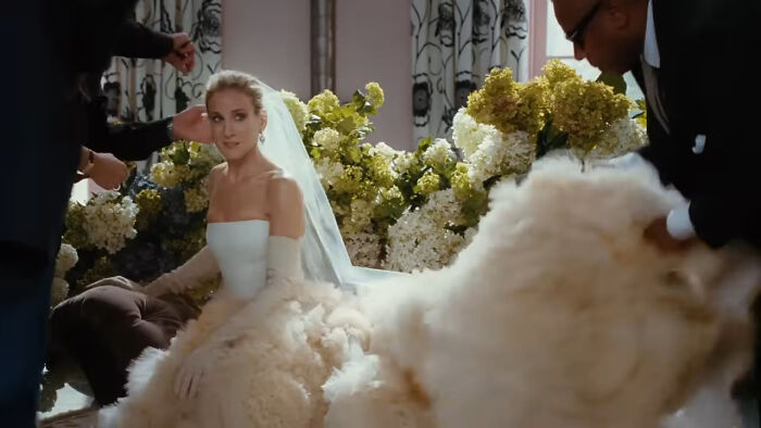Woman in an extravagant wedding dress and veil, surrounded by people adjusting her gown, set against floral decor.