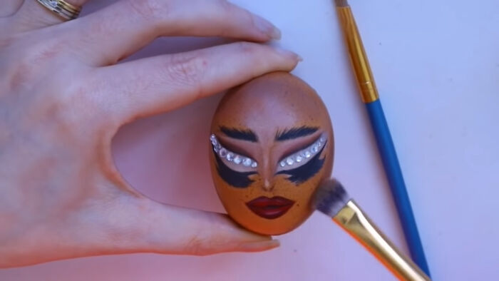 Decorated egg with dramatic makeup and gems, reflecting recent fashion trends people hate, held by a hand with a brush nearby.