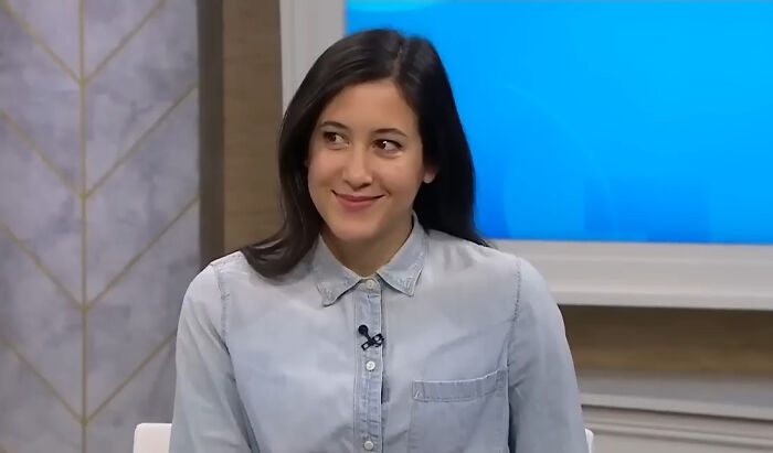 A woman in a light blue shirt, smiling during a pop culture discussion on a television set.