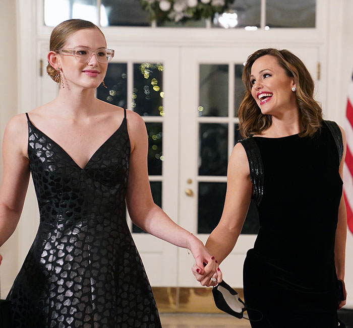 Two women, elegantly dressed, holding hands and smiling in a well-lit room.