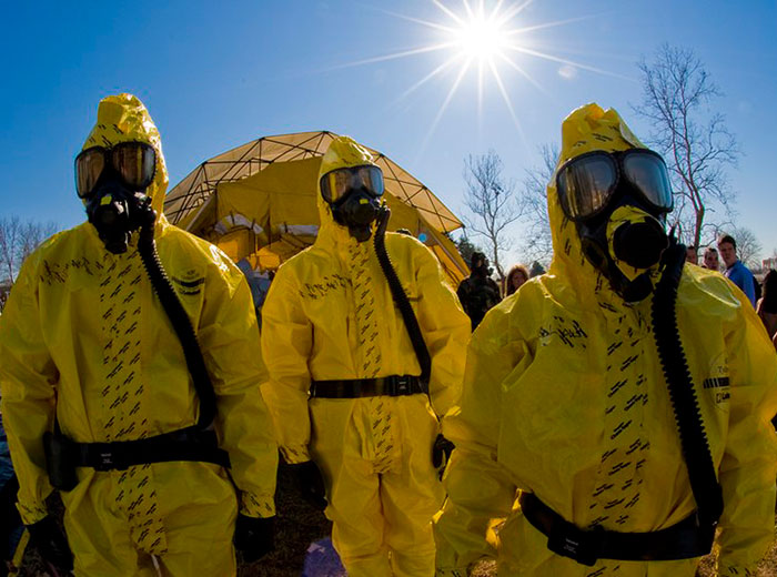 Experts in yellow hazmat suits investigate unknown illness site under the sun, with a tent and trees in the background. Experts in yellow hazmat suits investigate unknown illness site under the sun, with a tent and trees in the background.