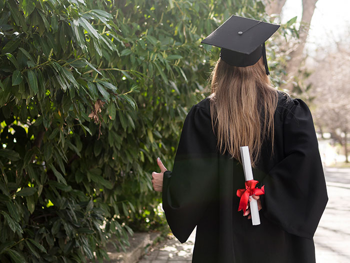 Graduate in cap and gown holding diploma with a red ribbon, representing things money can buy, showing a thumbs-up outdoors.