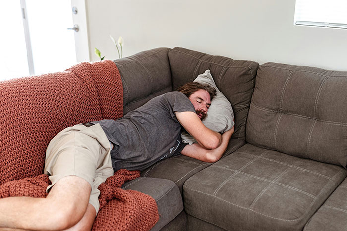 Man asleep on a couch displaying weaponized incompetence, wearing a gray shirt and beige shorts, surrounded by cozy blankets.