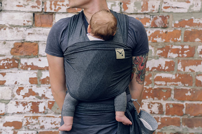 Man wearing baby carrier, illustrating weaponized incompetence, standing against a brick wall.