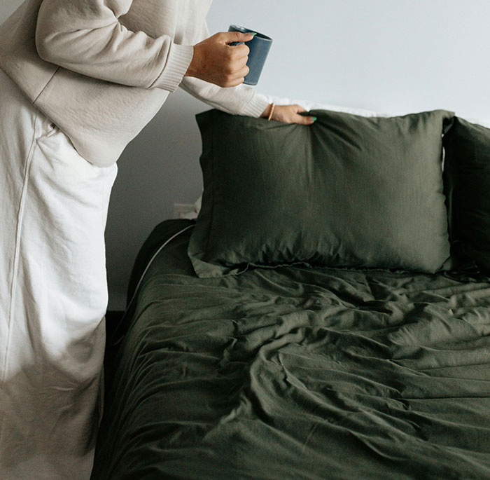 Person holding a cup while adjusting a green pillow on an unmade bed, illustrating weaponized incompetence in daily chores.