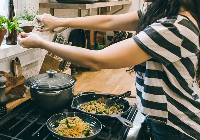A woman cooking pasta on a stovetop while trimming fresh herbs, illustrating daily tasks amid weaponized incompetence.
