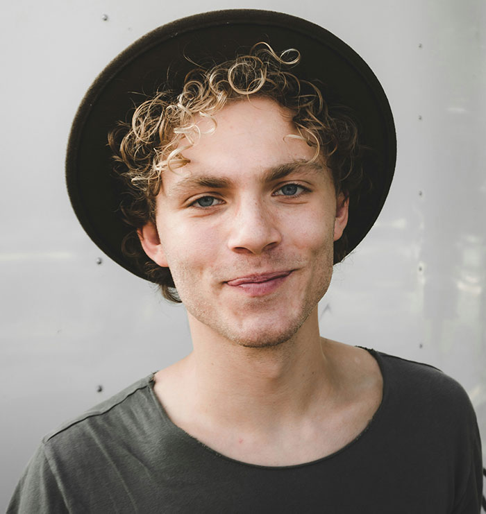 Young man with curly hair and a hat, smiling subtly; related to weaponized incompetence.