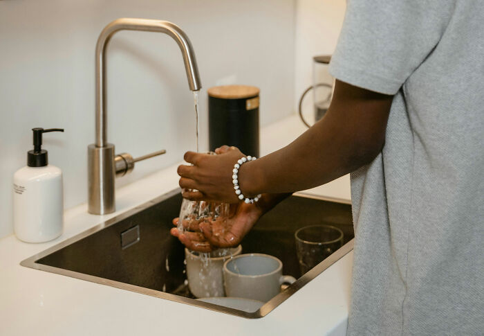 Woman washing dishes at a kitchen sink, wearing a bracelet, with water running over her hands.