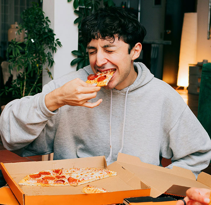 Man in a hoodie enjoying pizza from a box, representing guys appreciating pretty things too.