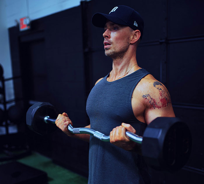 Man in gym lifting weights, wearing a cap and tank top, representing guys who like pretty things too.