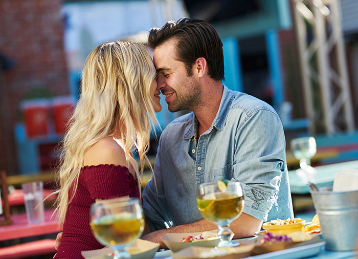 A couple sitting at an outdoor cafe, smiling and leaning towards each other with drinks on the table, enjoying Valentine's Day.