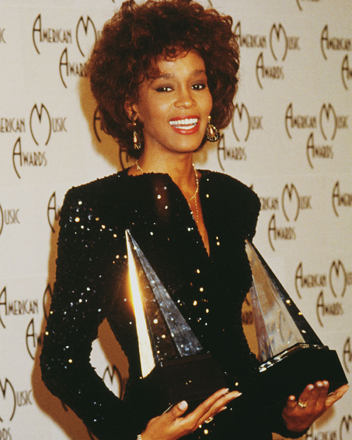 Celebrity at award show, holding two trophies, wearing a sparkling black dress, showing a wide smile.