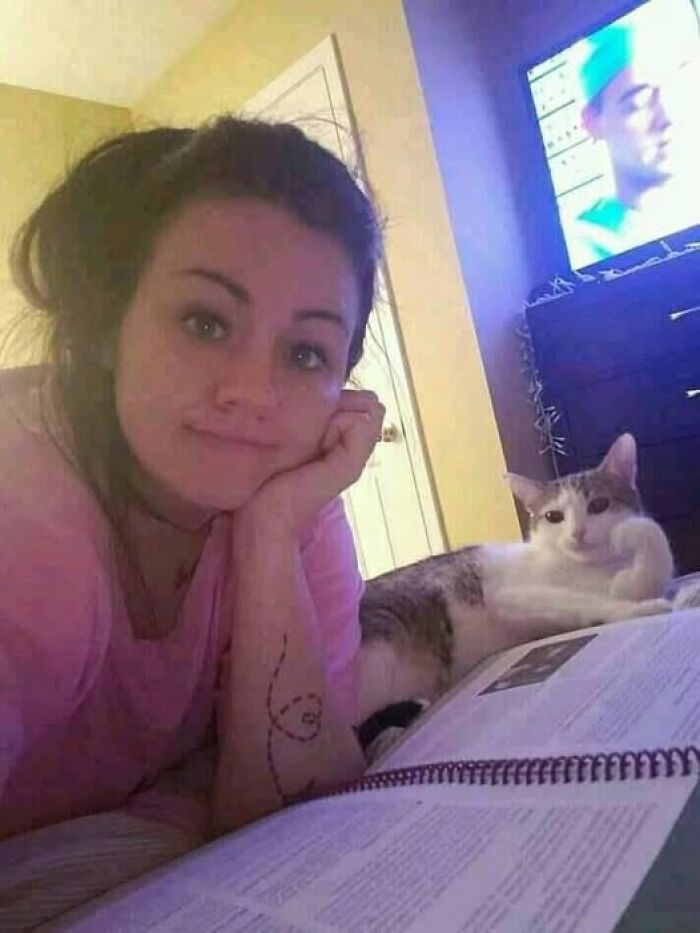 Woman and cat relaxing on a bed, with a book open and a TV in the background. Wholesome animal moment.