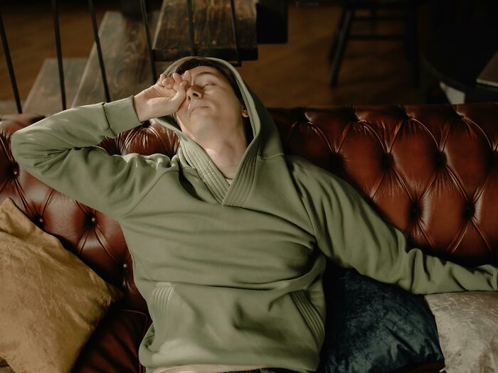 A man in a green hoodie resting on a brown couch, covering his face with his hand, possibly making old man noises. A man in a green hoodie resting on a brown couch, covering his face with his hand, possibly making old man noises.