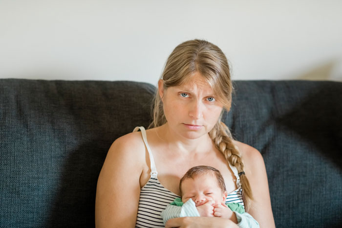 Woman sitting on a couch with a newborn baby, looking upset.