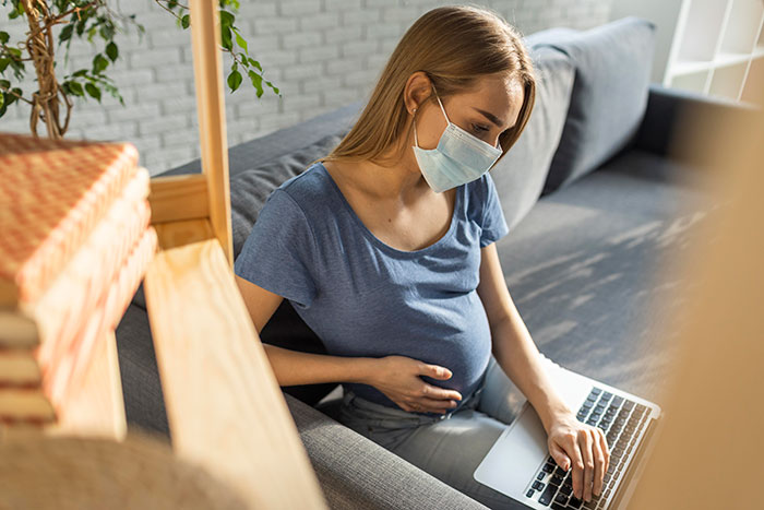 Pregnant woman in blue shirt and mask sitting on a couch, using a laptop.