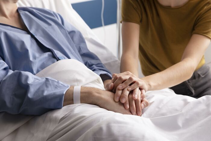 Person in hospital bed being lovingly supported by another holding their hands.