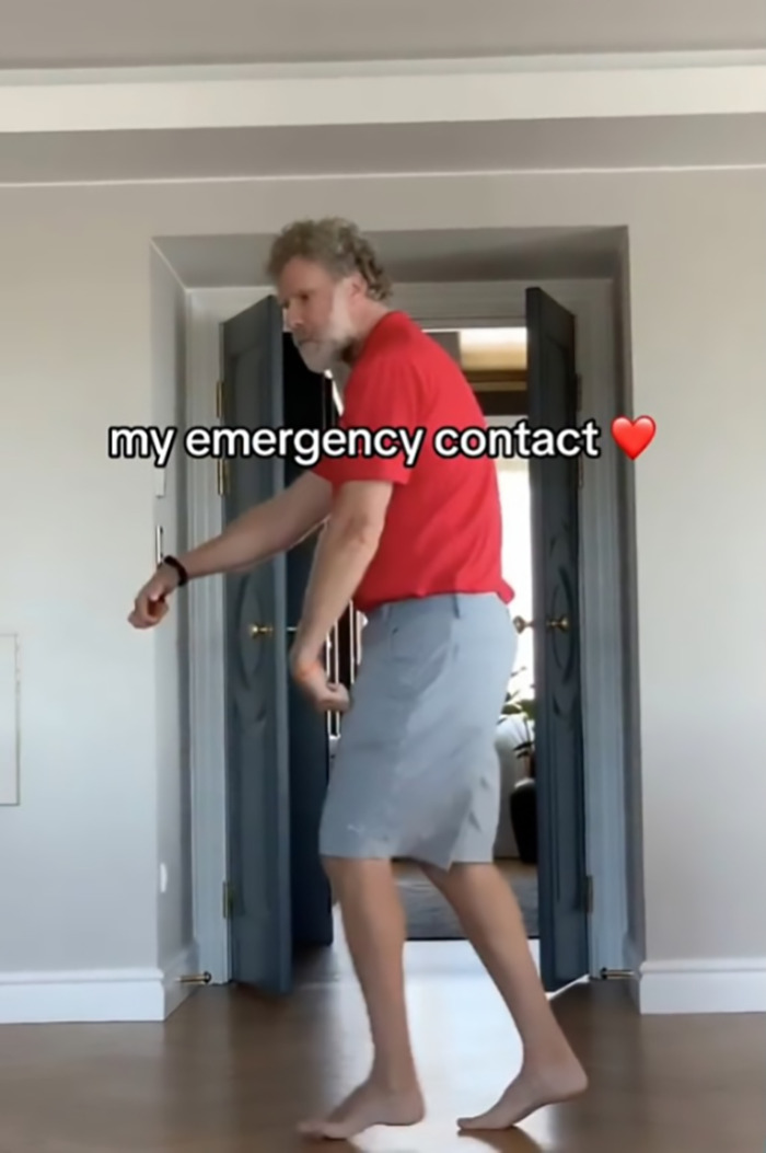 Will Ferrell in a red shirt and gray shorts dancing humorously in a room, captioned "my emergency contact" with a heart emoji.