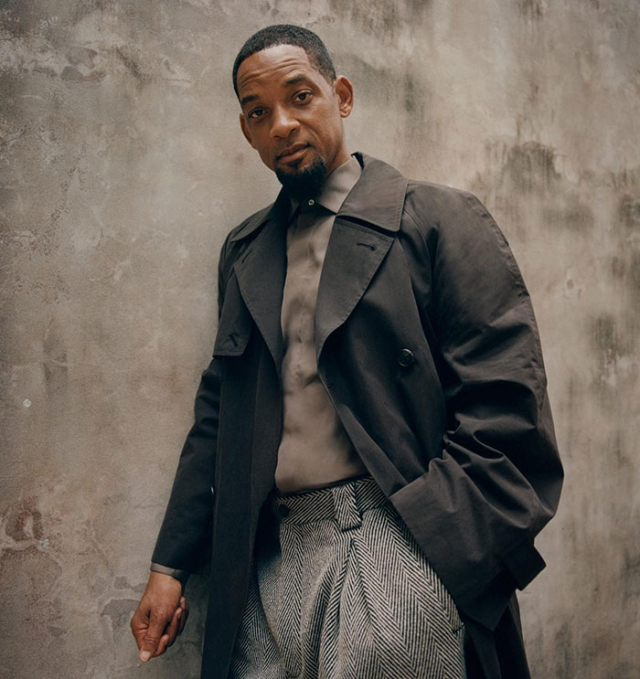 Man in a dark coat and patterned trousers stands against a concrete wall, exuding "creepy old man vibes.
