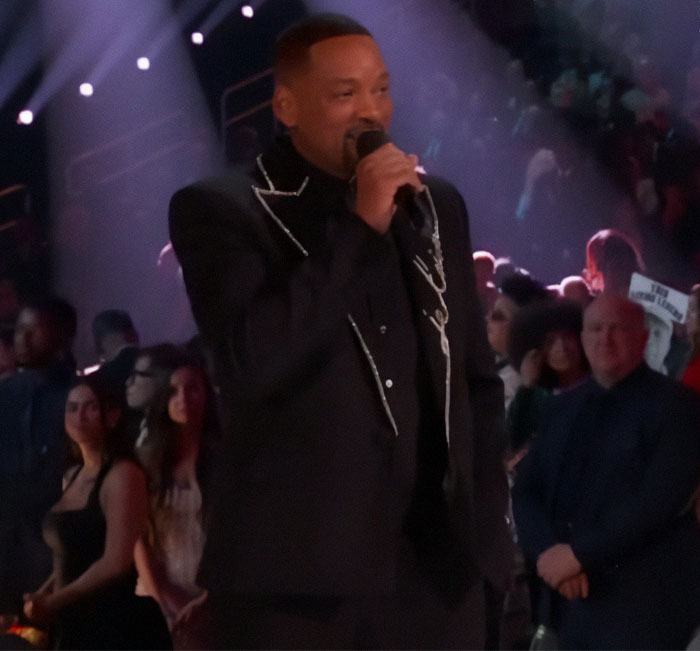 "Man in a black suit on stage, holding a microphone at an event, with audience members in the background. "Man in a black suit on stage, holding a microphone at an event, with audience members in the background.