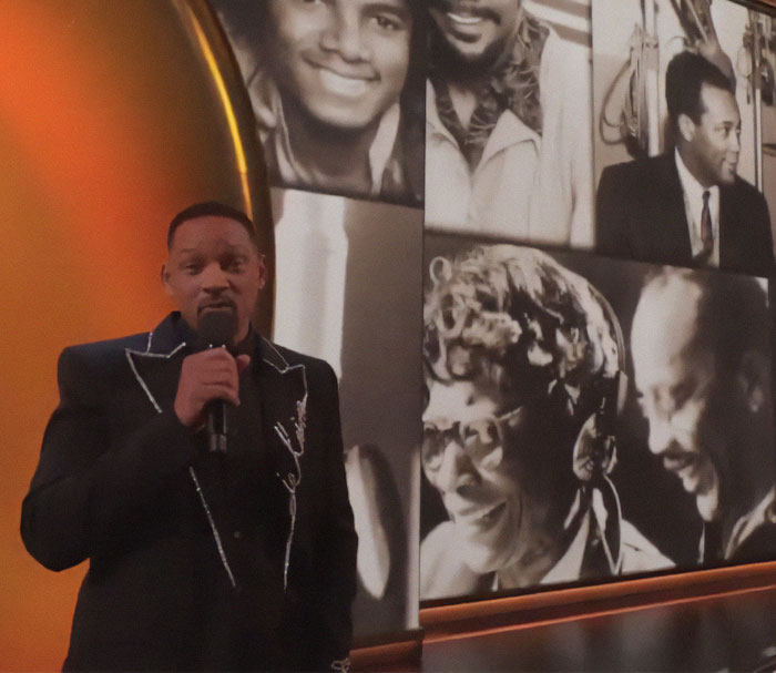 Man in black suit speaks at event, standing near a collage of black and white photos, related to Quincy Jones tribute. Man in black suit speaks at event, standing near a collage of black and white photos, related to Quincy Jones tribute.