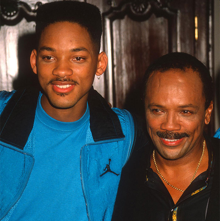 "Two smiling men embracing indoors, one wearing a blue jacket, in a tribute setting. "Two smiling men embracing indoors, one wearing a blue jacket, in a tribute setting.