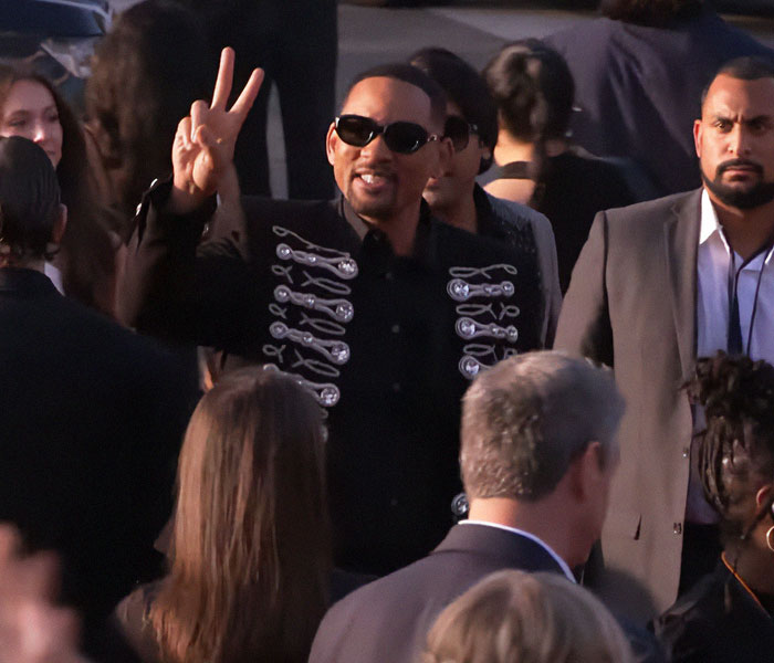 Person at the 2025 Grammy Awards, wearing sunglasses and a stylish black jacket, making a peace sign in a crowded scene.