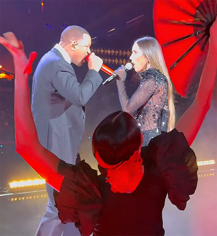 Man and female singer performing onstage with vibrant red lighting and an umbrella, conveying "creepy old man vibes.