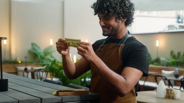 Young barista in an apron examining a dollar bill, reflecting on life lessons in a cozy cafe setting.