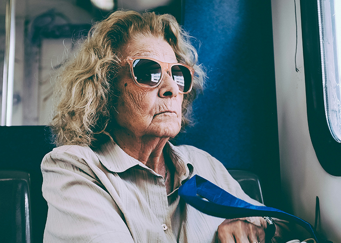 Elderly woman with sunglasses, sitting indoors, reflecting on son's girlfriend's house rule.